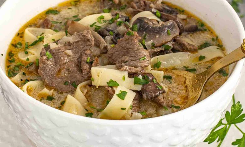 A close-up shot of a bowl of Beef Stroganoff Soup served in a textured white bowl on a matching plate. The creamy, golden broth is packed with tender slices of beef, wide egg noodles, and earthy mushrooms, all garnished with freshly chopped parsley.