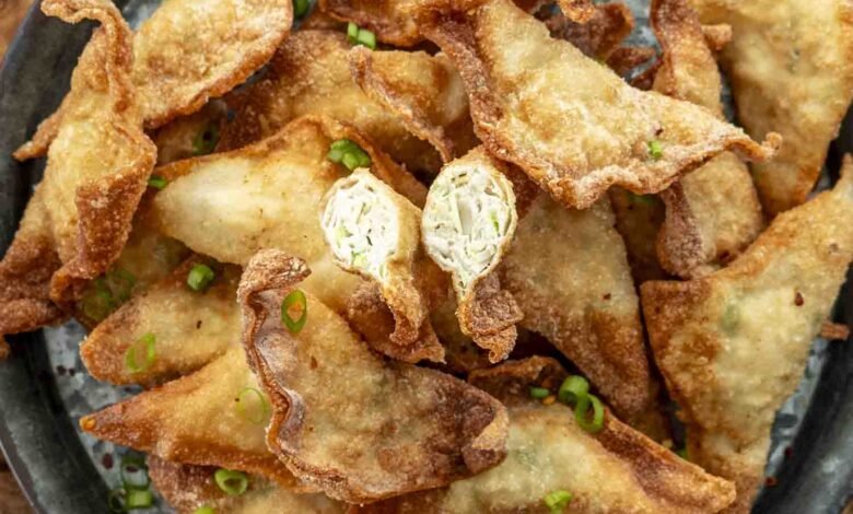 A black metal serving tray filled with crispy, golden-brown chicken fried wontons, garnished with finely chopped green onions. One wonton is cut open, revealing a juicy, well-seasoned chicken and cabbage filling. A small bowl of dipping sauce with chili flakes sits in the background, adding a rich, savory contrast to the crunchy wontons.