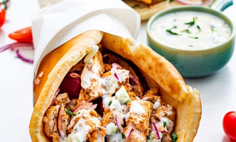 a chicken gyro wrapped in paper and topped with lots of tzatziki sauce with another gyro in the background