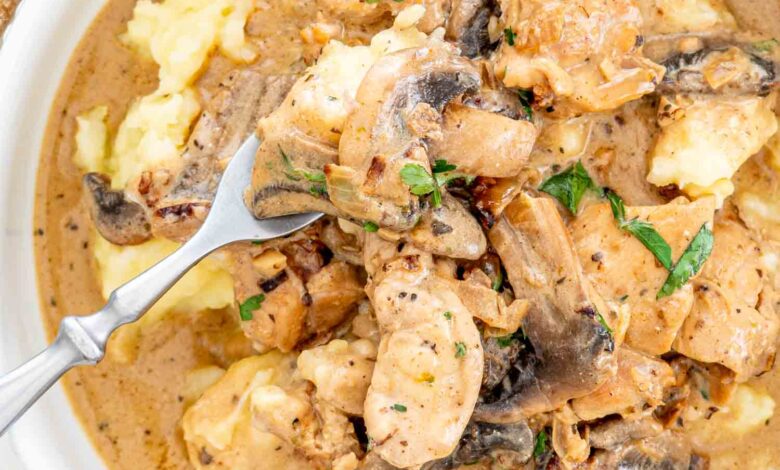 A bowl of chicken stroganoff with mushrooms atop smooth mashed potatoes, sprinkled with herbs.