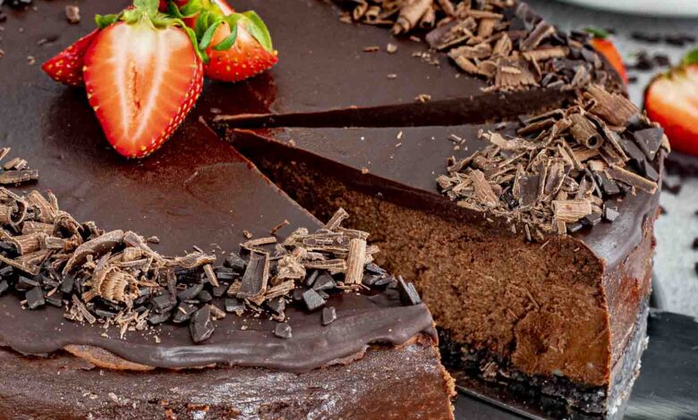 A rich and creamy chocolate cheesecake with a glossy ganache topping, garnished with fresh strawberries and chocolate shavings. A slice is being lifted, revealing the velvety chocolate filling and a dark, crumbly chocolate cookie crust.