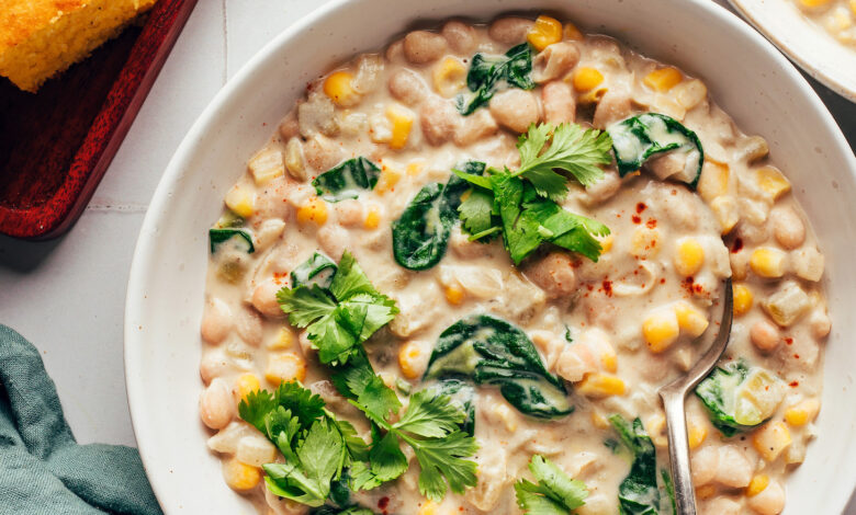 Bowls of creamy vegan white bean chili next to cayenne, cilantro, and cornbread