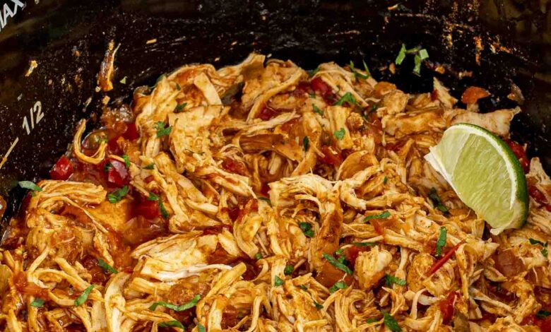 Shredded Slow Cooker Mexican Chicken in a black slow cooker, garnished with fresh cilantro and lime wedges.